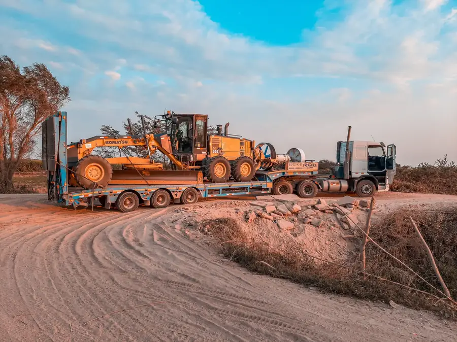 Grader chained down on a three-axle low loader on a dusty dirt road