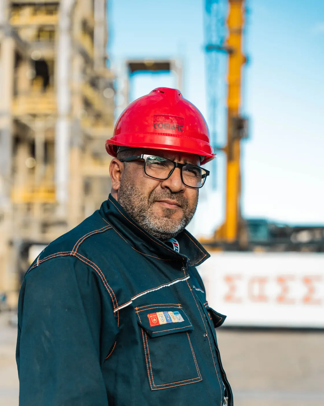 Crew supervisor in a red hard hat standing before a crane and plant works