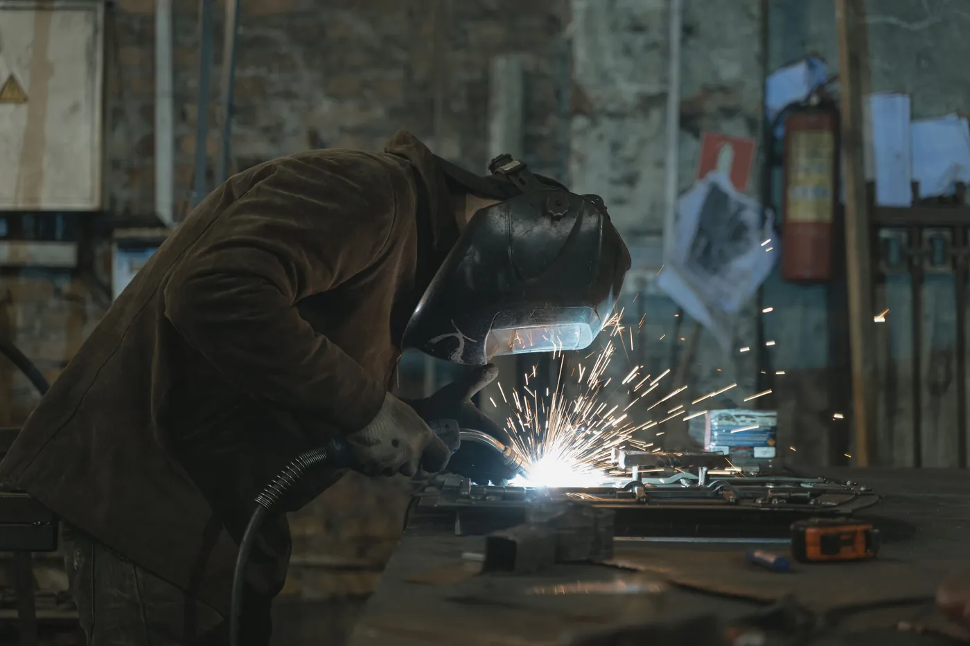 Welder working at a bench in a dark fabrication workshop, sparks flying